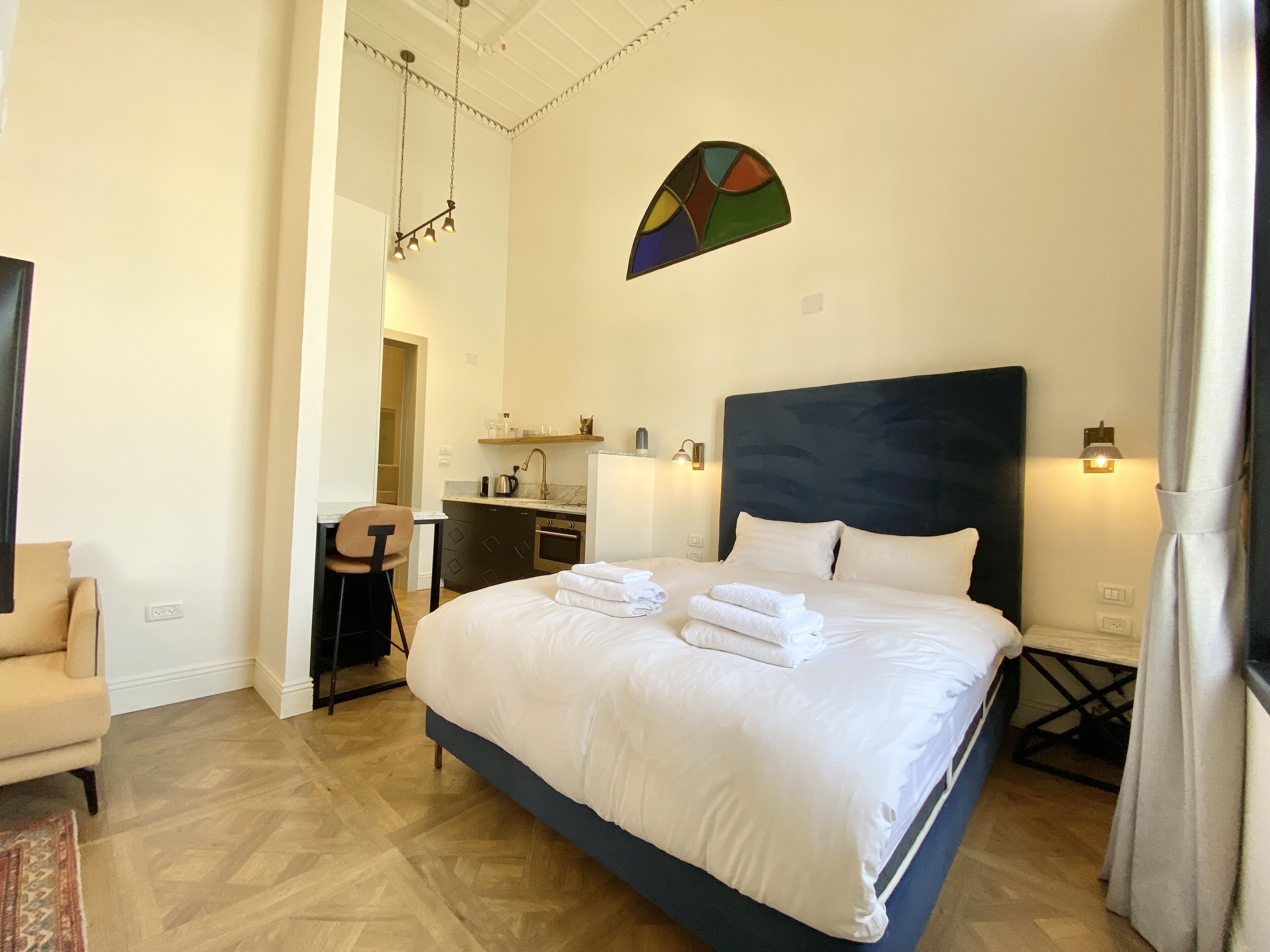 A hotel room with a large bed, a desk, a chair, a small kitchenette, and a bathroom.