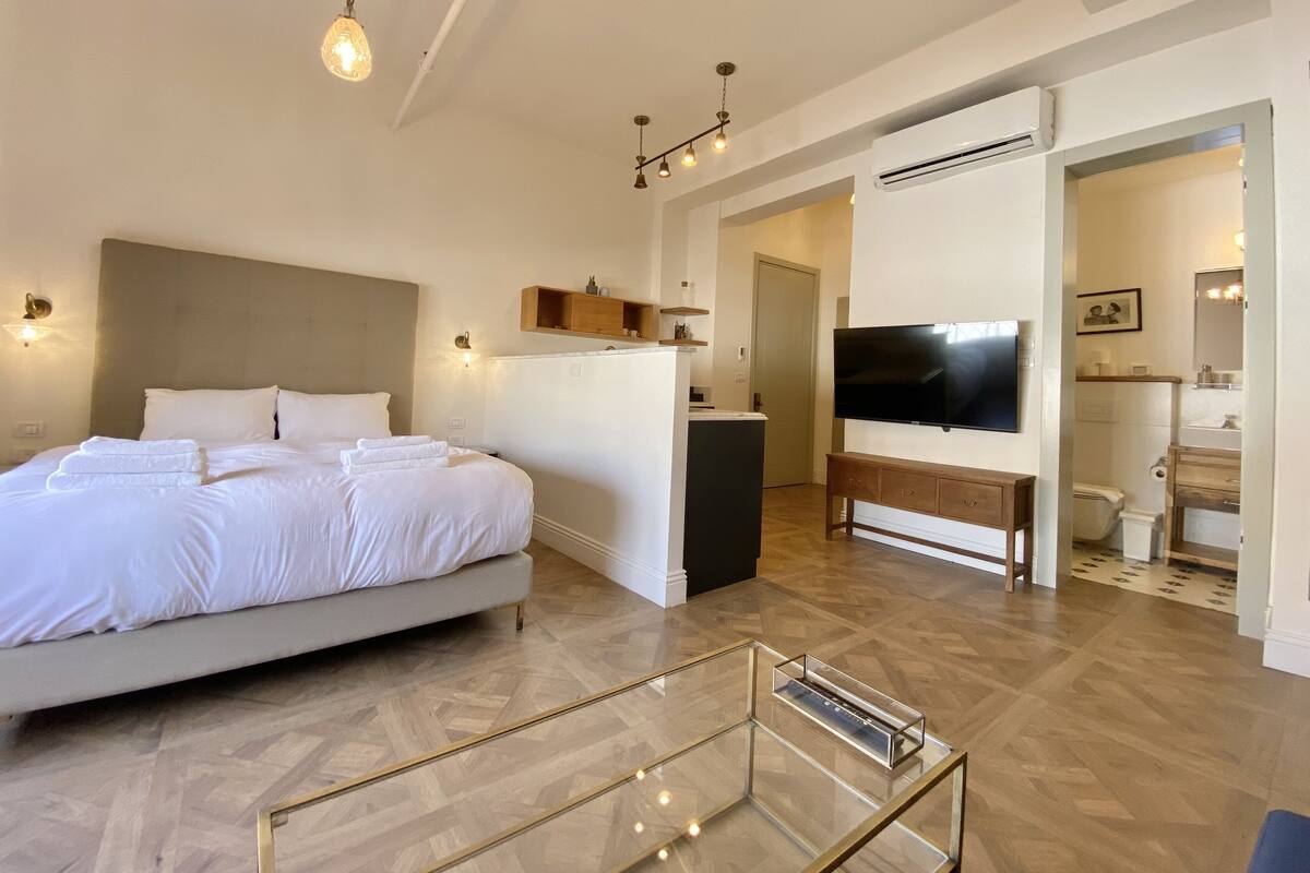 A modern hotel room with a large bed, a TV, a small desk, and a glass coffee table.