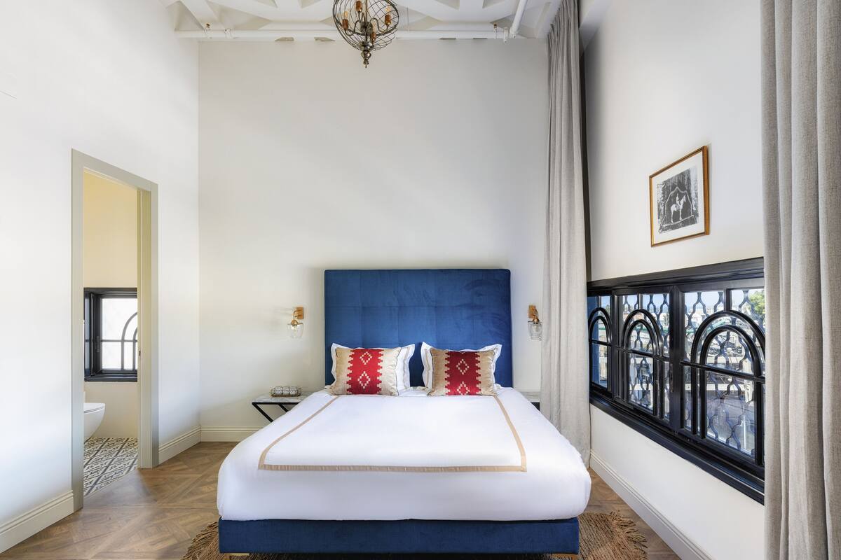 A bedroom with a large bed, a chandelier, a window with a view, and a framed picture on the wall.