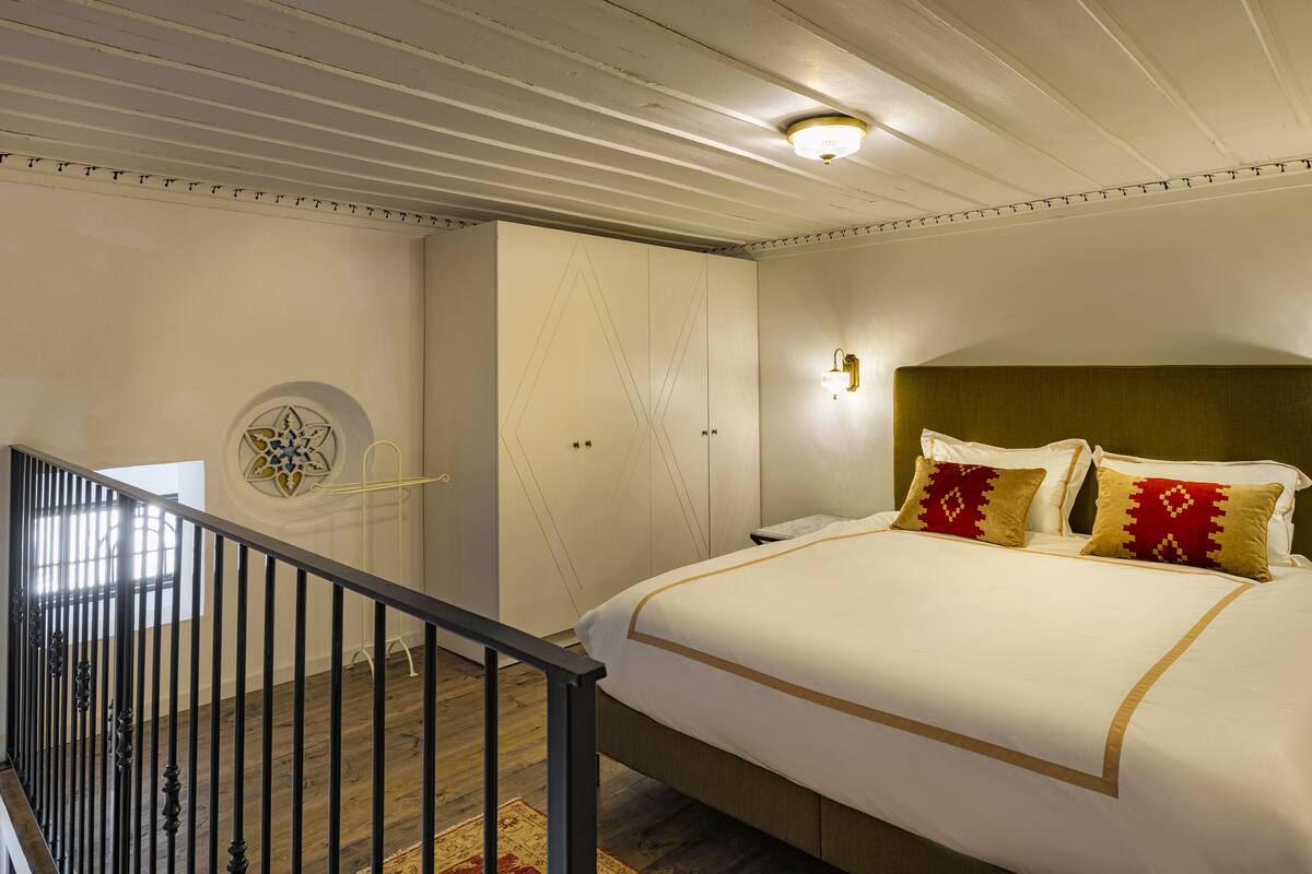 A hotel room with a large bed, a headboard, two wall sconces, a ceiling light, a small table, a nightstand, a window with blinds, a door, and a staircase with a railing.