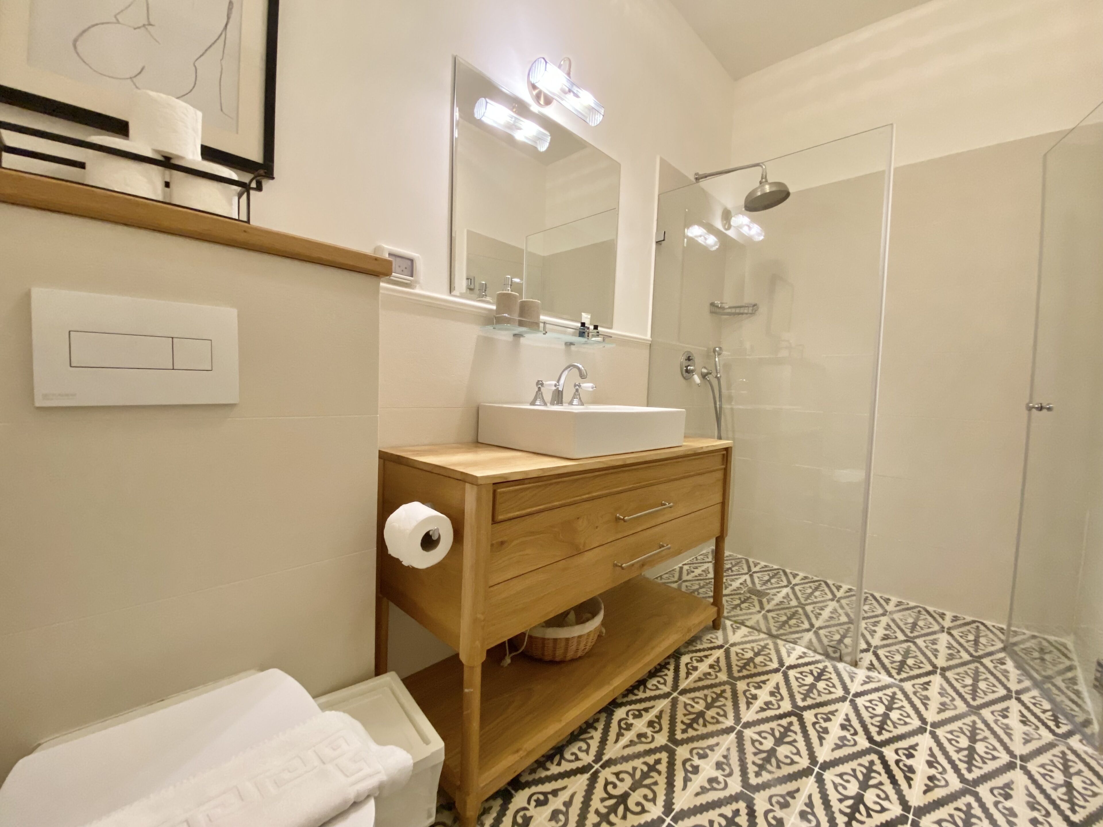 A modern bathroom with a wooden vanity, a large mirror, a shower, and patterned floor tiles.