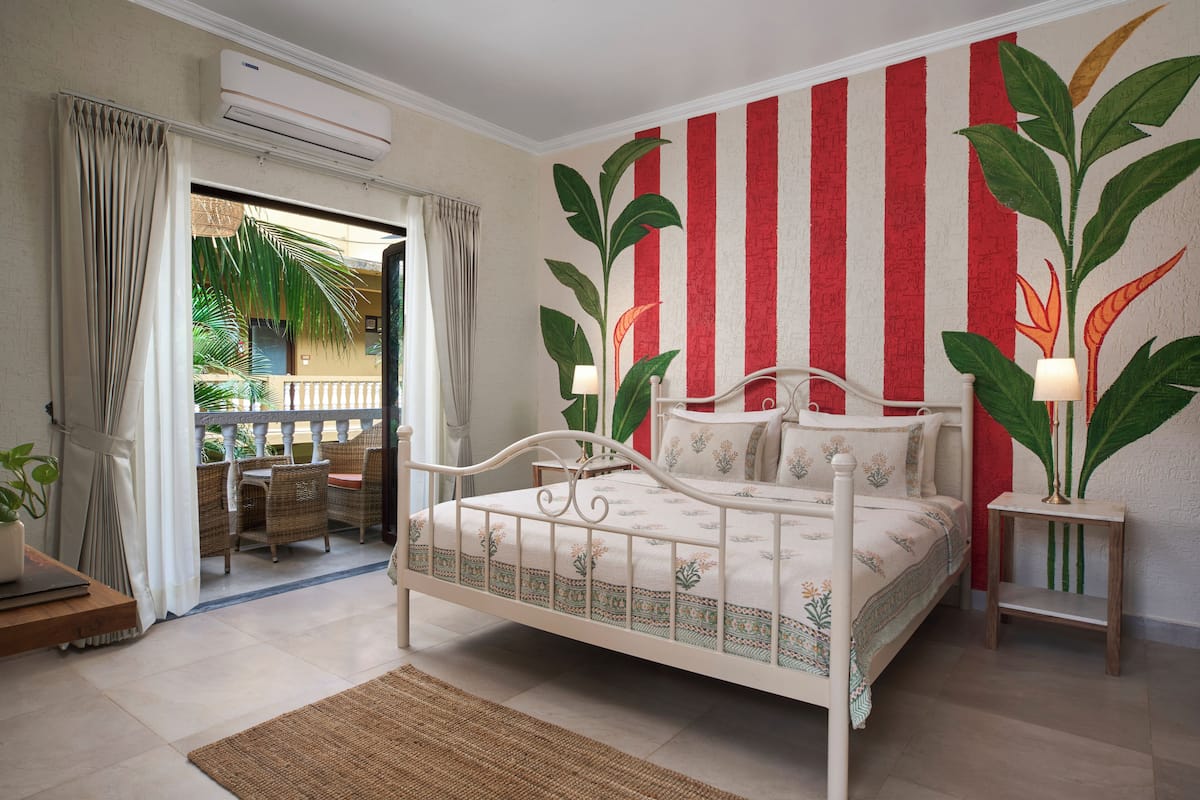 A bedroom with a bed, a small table, a lamp, and a view of the outside.
