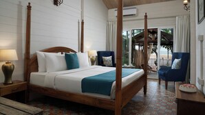 A four-poster bed with a wooden frame, white bedding, and teal throw pillows. There are two bedside tables with lamps, a blue armchair, and a small table with a bowl of fruit. The room has a tiled floor and a view of the outdoors through a glass door. - Larisa Resort, Morjim (Morjim)