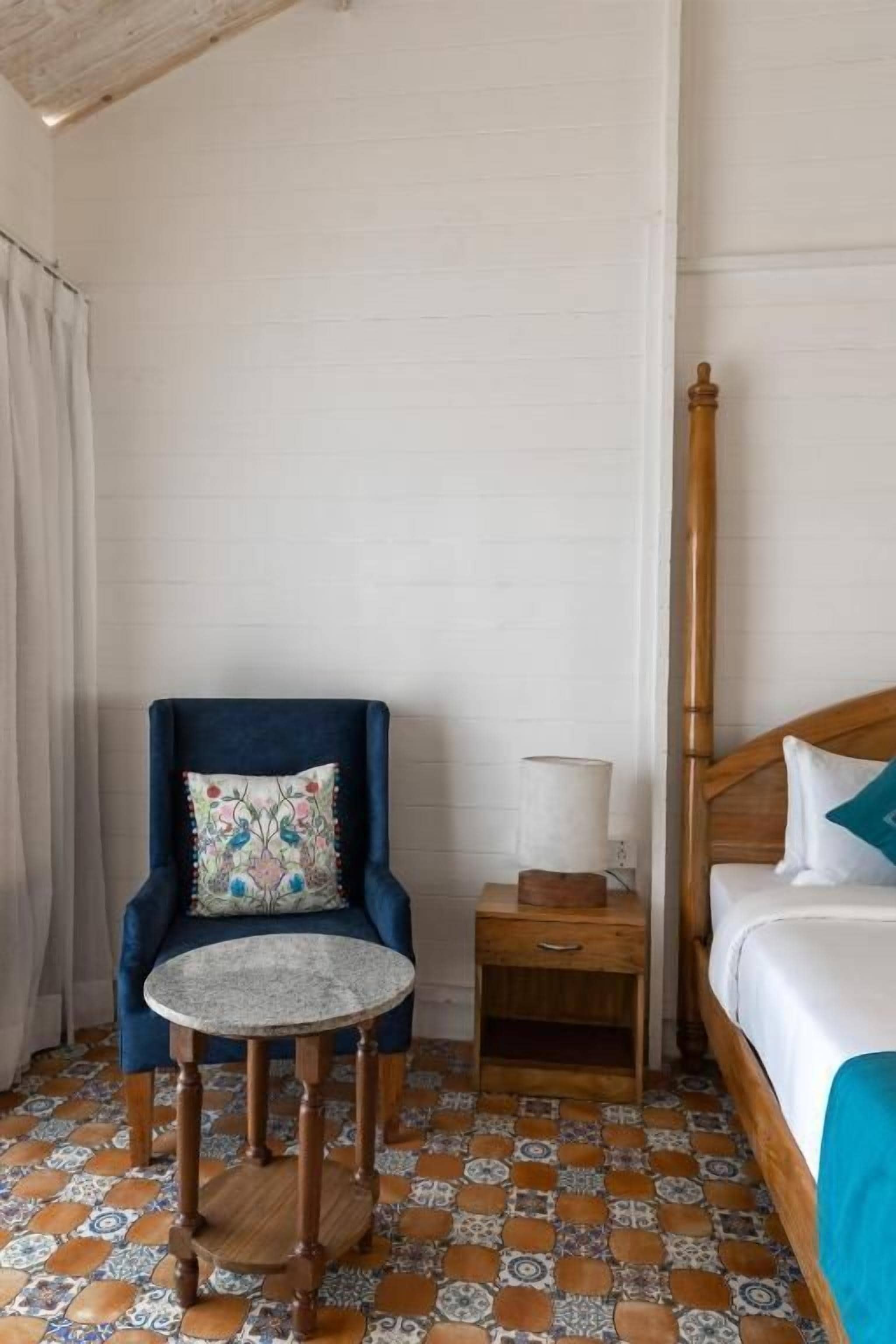 A room with a blue armchair, a small round table, a wooden nightstand, and a bed with a wooden headboard.