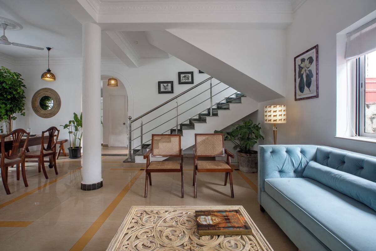 A spacious living room with a blue sofa, wooden chairs, a dining table with chairs, a staircase, and a large mirror on the wall.