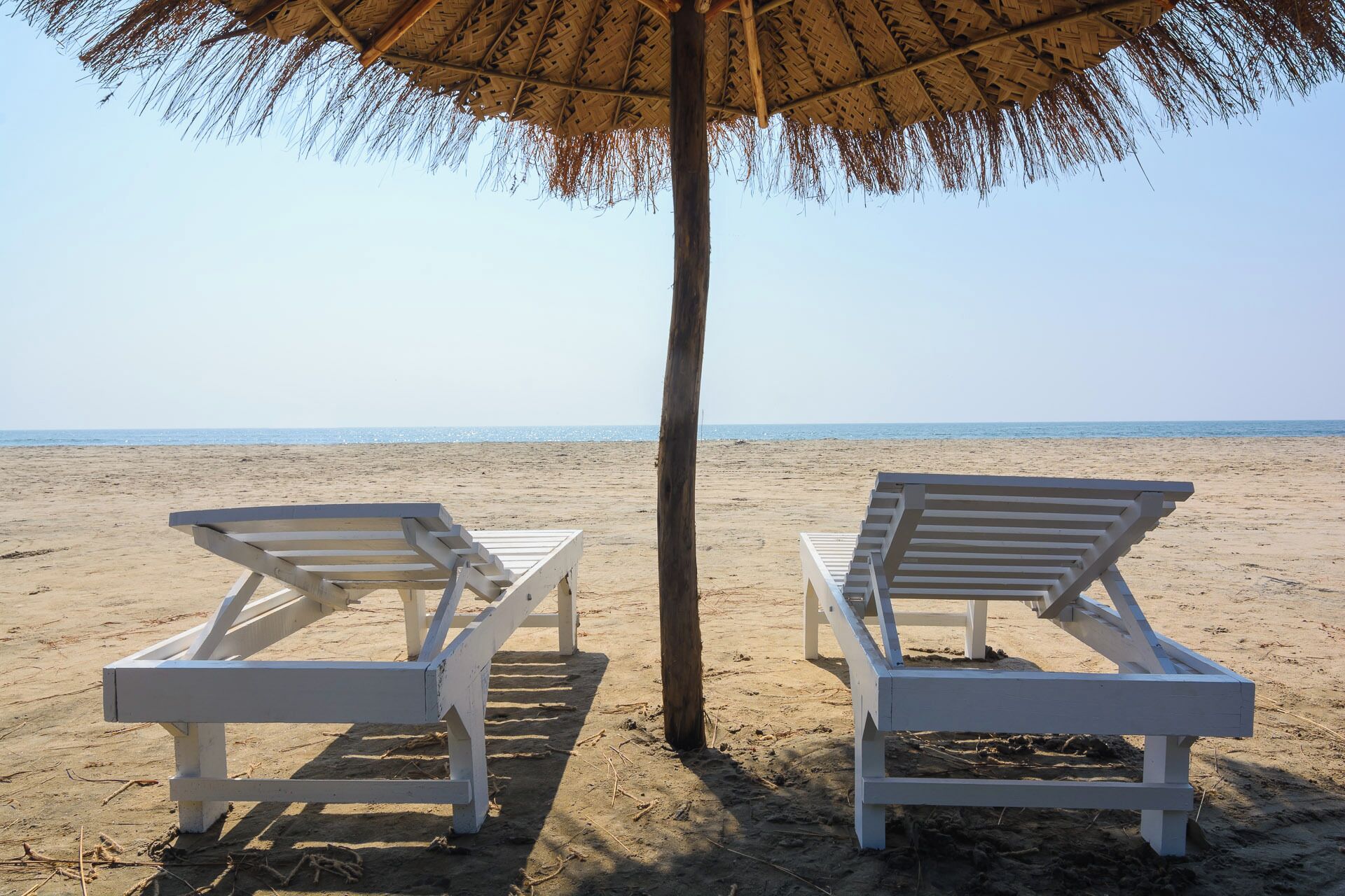 Two white lounge chairs under a thatched umbrella on a sandy beach with a clear view of the ocean.