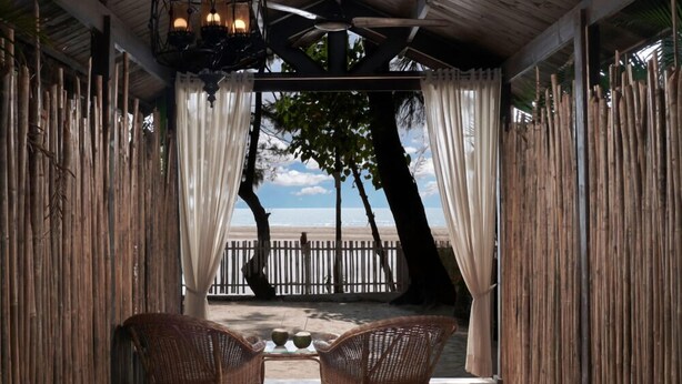 A covered outdoor seating area with wicker chairs, a large planter with red flowers, and a view of the beach. - Larisa Resort, Morjim (Morjim)