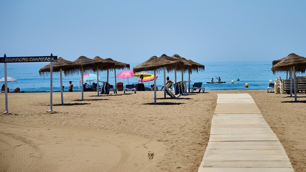 Une plage avec des parasols en chaume, des chaises longues et une passerelle en bois menant à l’eau.