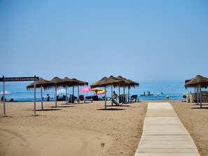 Ein Strand mit reetgedeckten Sonnenschirmen, Liegestühlen und einer Holzpromenade, die zum Wasser führt.