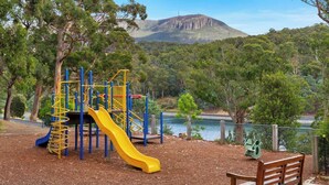 A playground with a yellow slide and climbing structures, a wooden bench, and a lake with a mountain in the background. - Explore South Hobart Village from an 1855 Cottage (Dynnyrne)