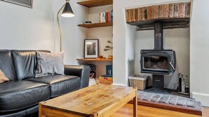 A living room with a black leather sofa, a wooden coffee table, a black lamp, a bookshelf, and a wood-burning stove. - Explore South Hobart Village from an 1855 Cottage (Dynnyrne)