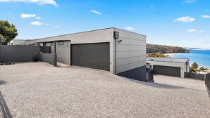 A modern building with a large garage door, a gravel driveway, and a view of the ocean. - Beachside Luxury Villa Expansive Panoramic Vistas (Kingston Beach)