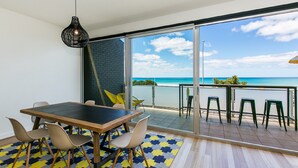 A modern interior with a dining area, a large sliding glass door leading to a balcony with a view of the ocean. - On The Beach (Lorne)