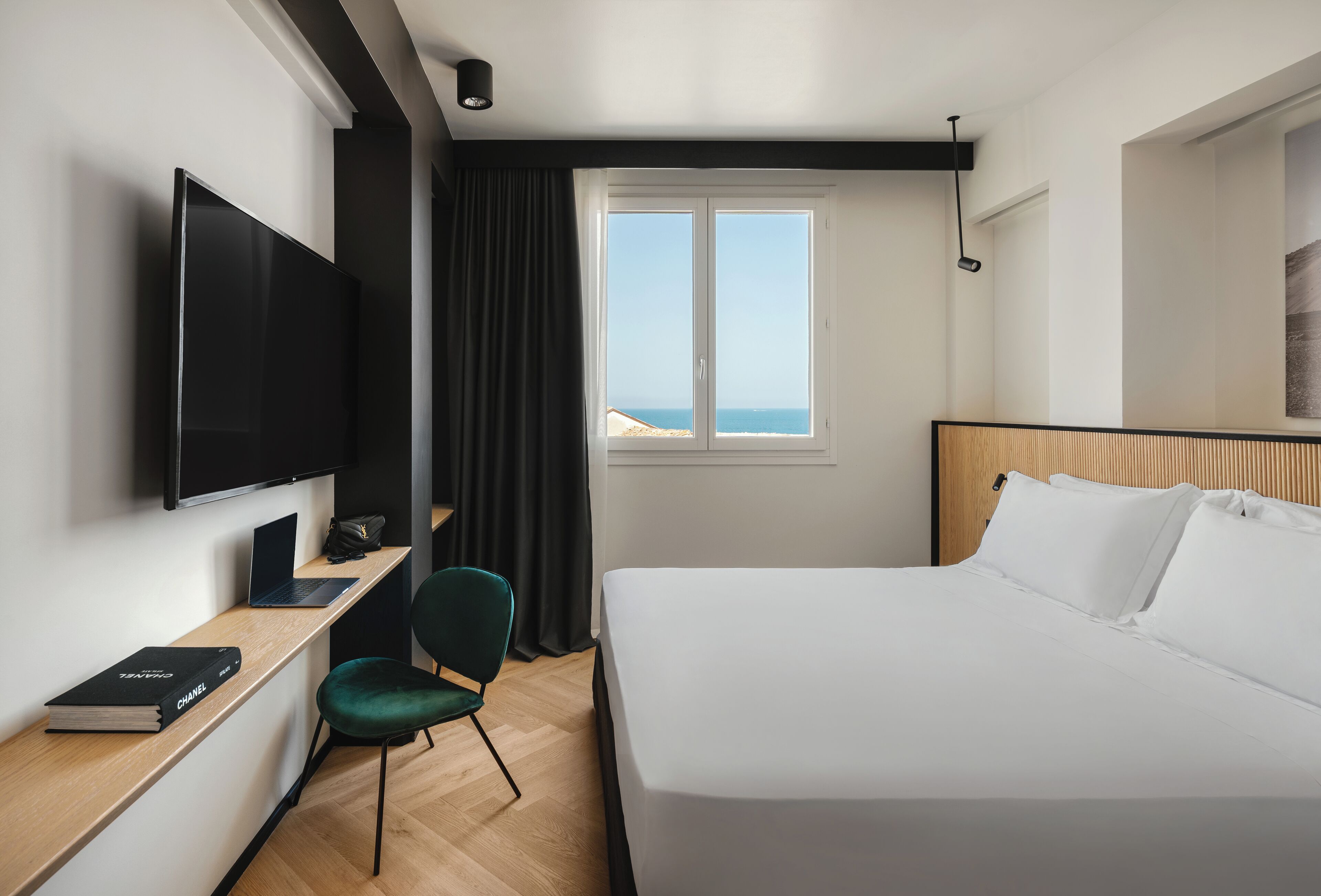 A hotel room with a bed, a desk with a laptop, a television, a chair, and a window with a view of the sea.