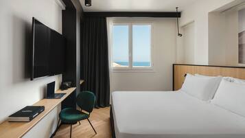 A hotel room with a bed, a desk with a laptop, a television, a chair, and a window with a view of the sea.
