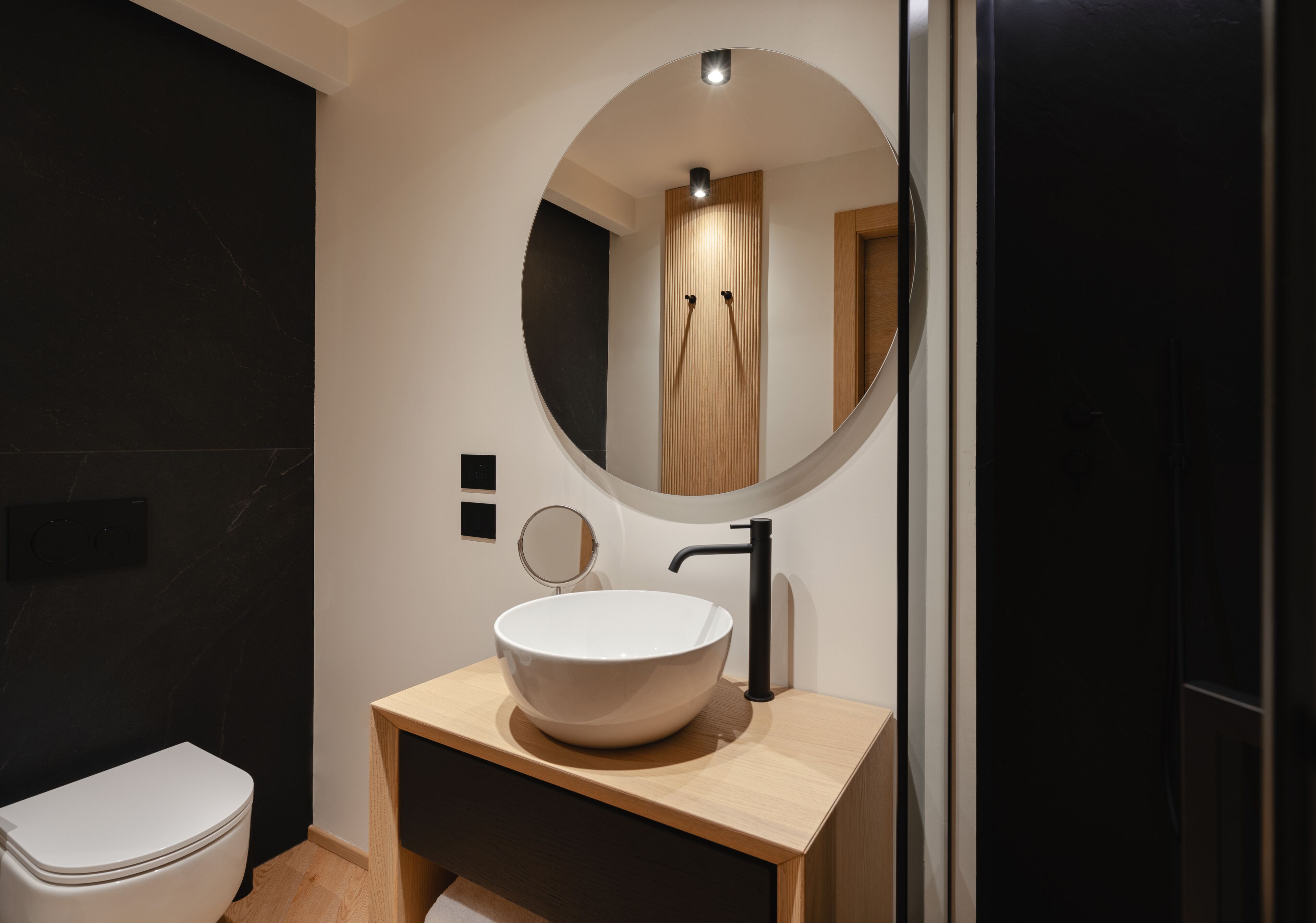 Ein modernes Badezimmer mit einer weißen Toilette, einem Holzwaschtisch, einem großen runden Spiegel und einem schwarzen Wasserhahn.