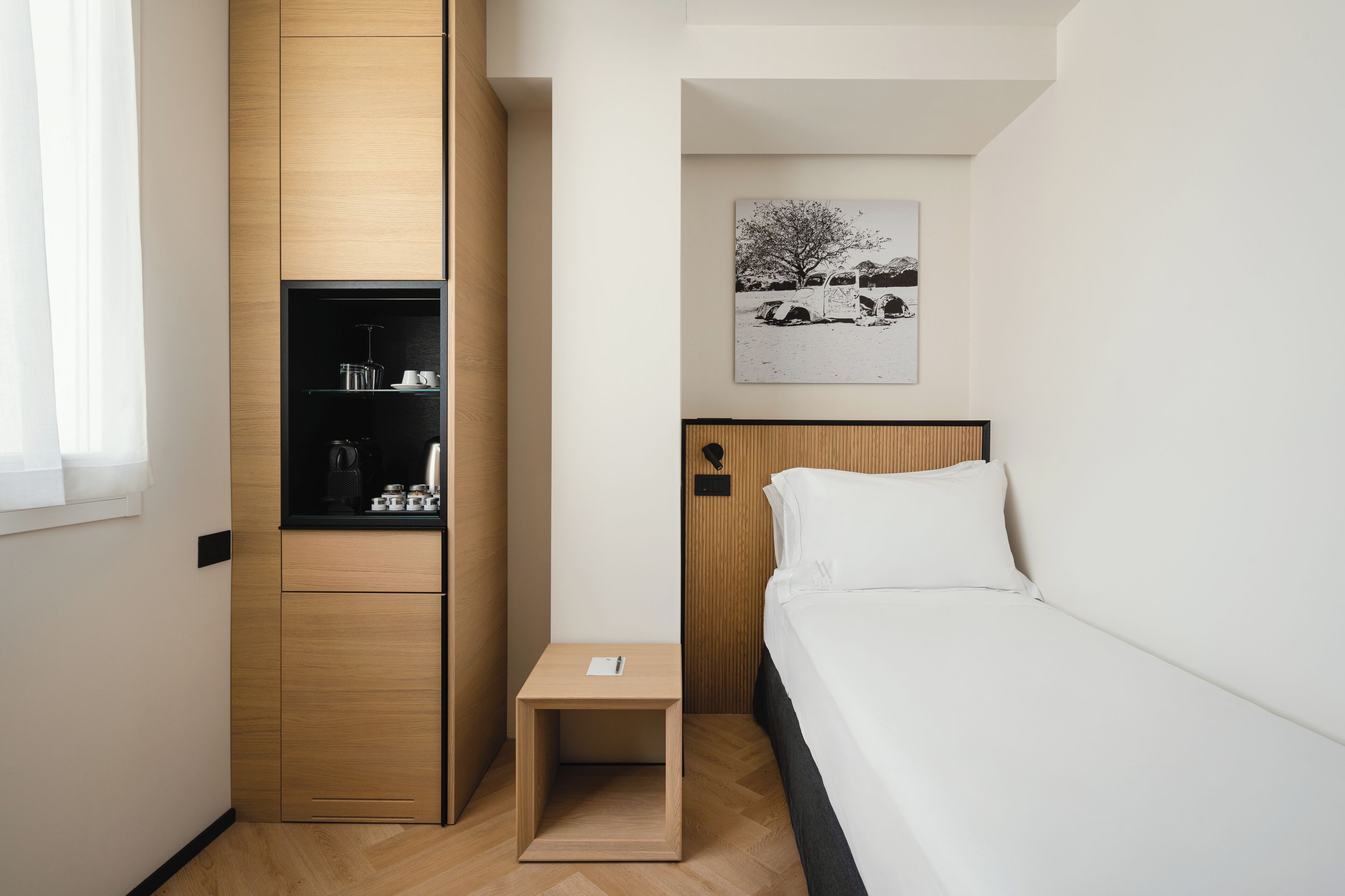 A compact hotel room with a bed, a small bedside table, a wardrobe, and a wall-mounted artwork.