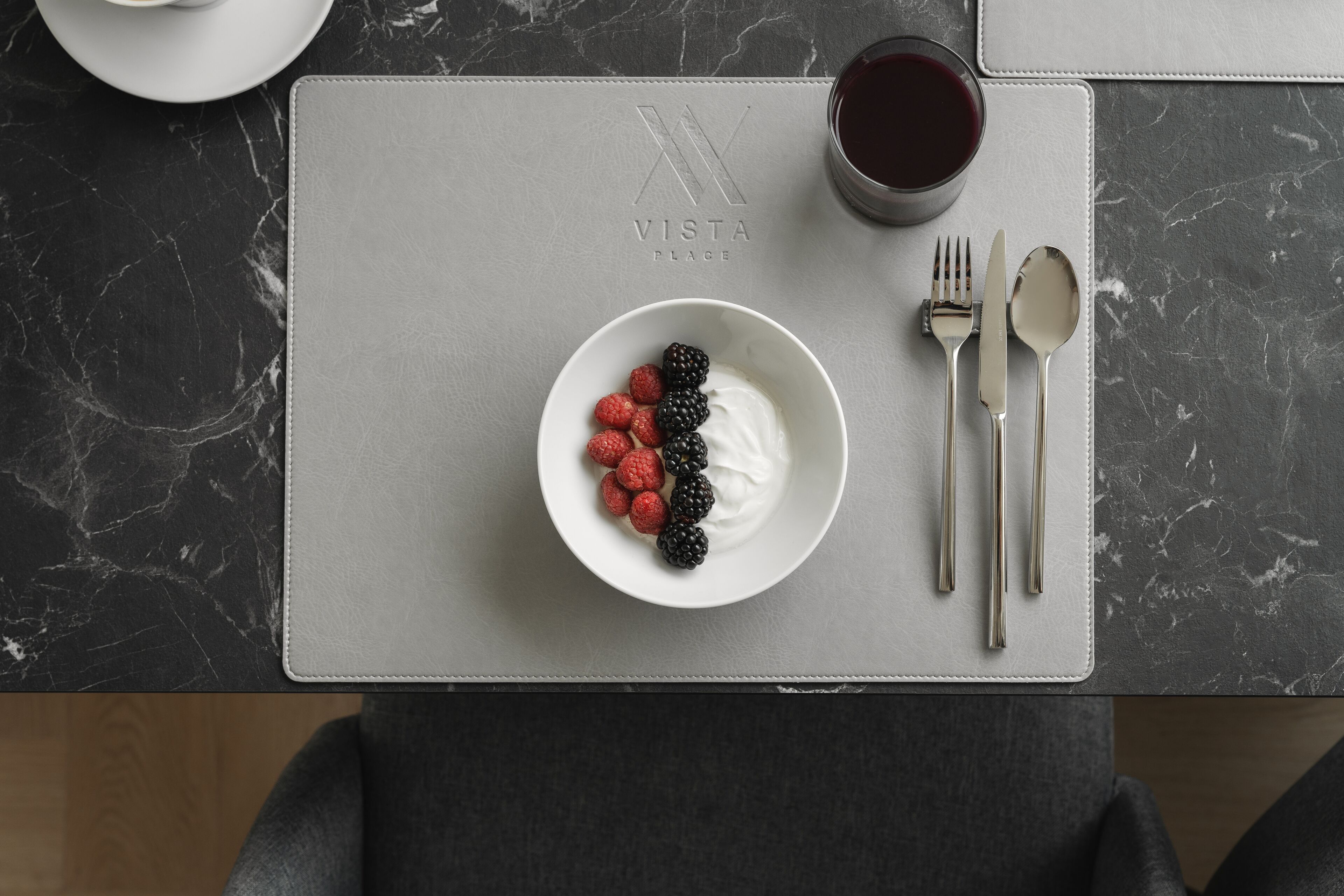 A table with a white placemat, a bowl of yogurt with berries, cutlery, and a glass of red drink.