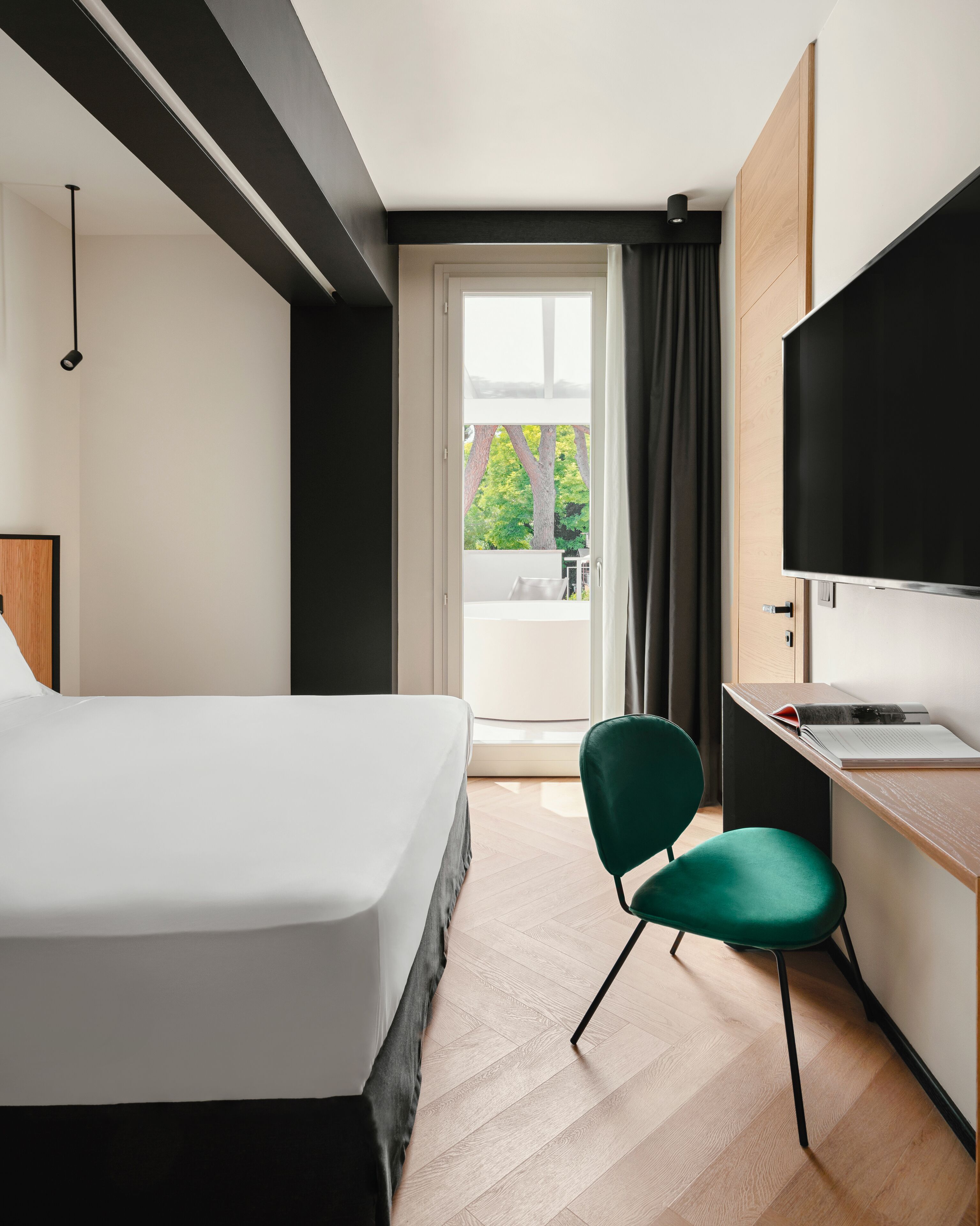 Una habitación de hotel moderna con cama, escritorio, silla, televisor y vista a los árboles a través de una ventana.