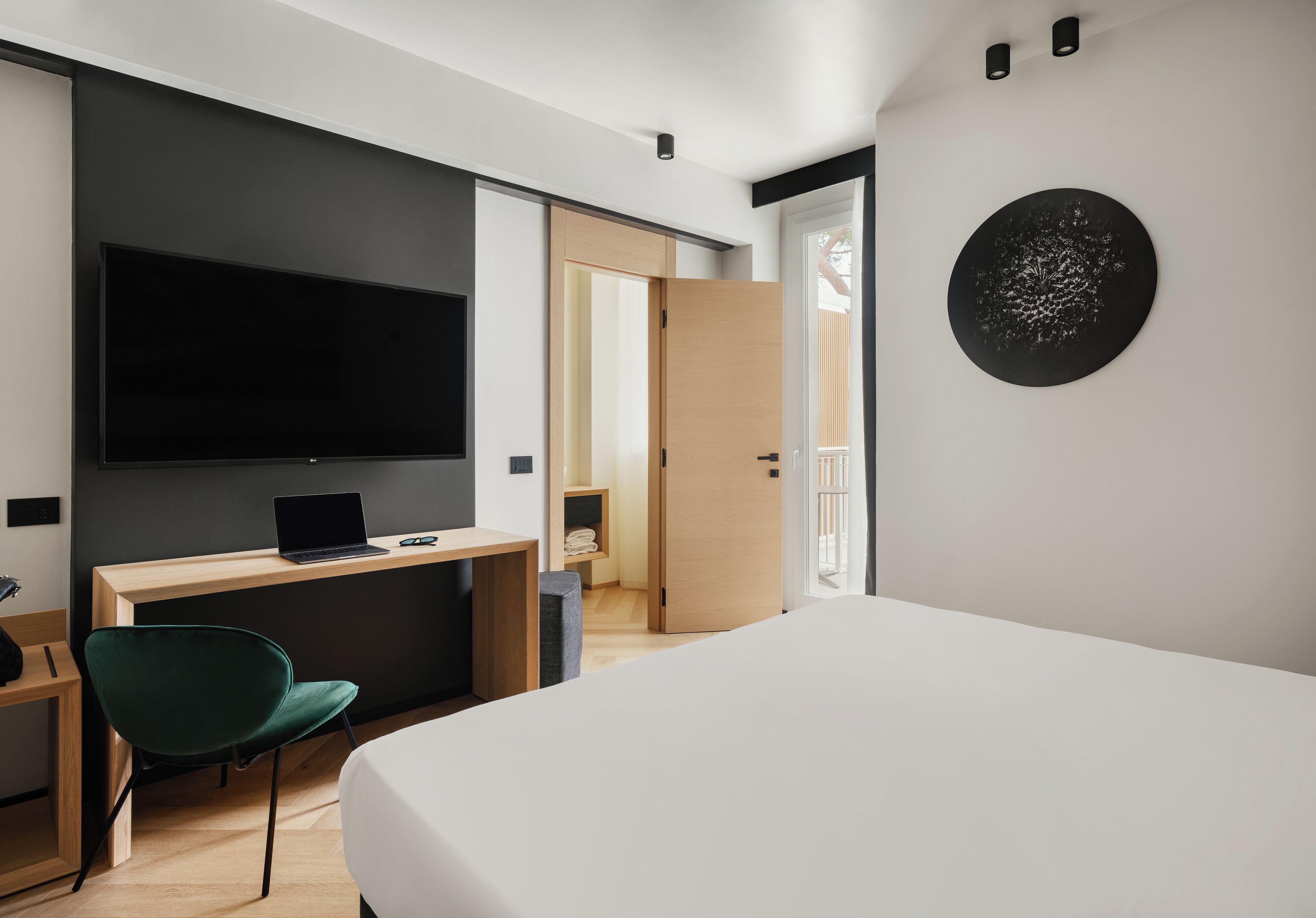 Ein modernes Hotelzimmer mit Bett, Schreibtisch mit Laptop, grünem Sessel, Flachbildfernseher und einem runden Kunstwerk an der Wand.