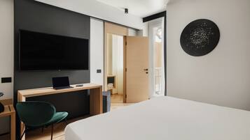 Ein modernes Hotelzimmer mit Bett, Schreibtisch mit Laptop, grünem Sessel, Flachbildfernseher und einem runden Kunstwerk an der Wand.