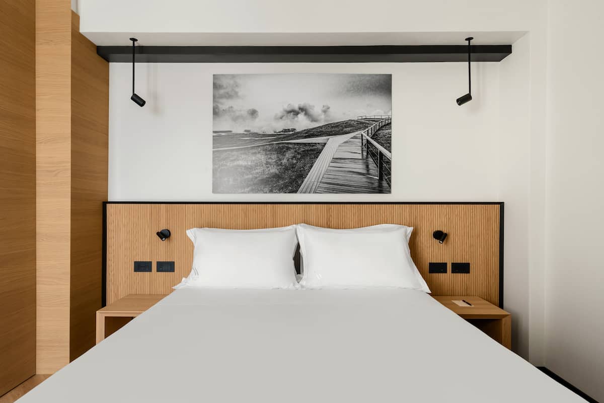 A modern hotel room with a wooden headboard, a large bed, two bedside tables, and a framed black and white photograph above the bed.