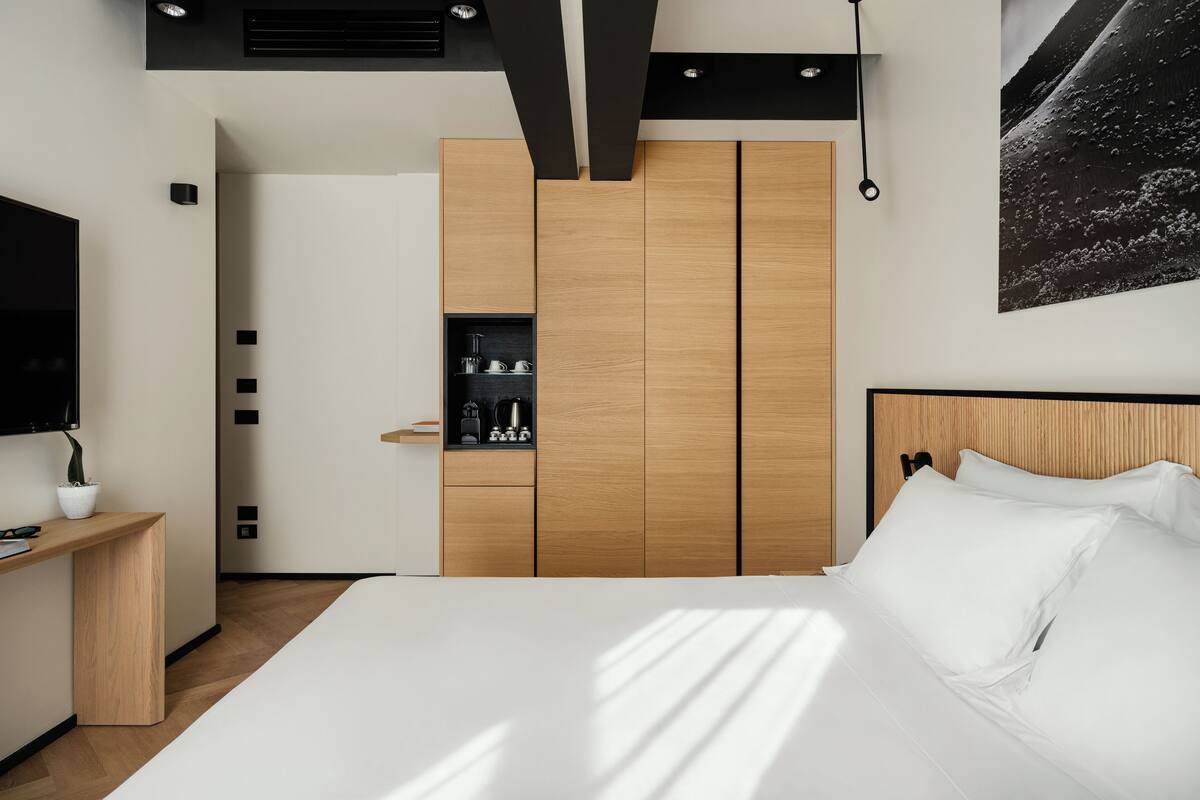 A modern bedroom with a large bed, a wooden wardrobe, a small shelf with toiletries, and a mounted TV.