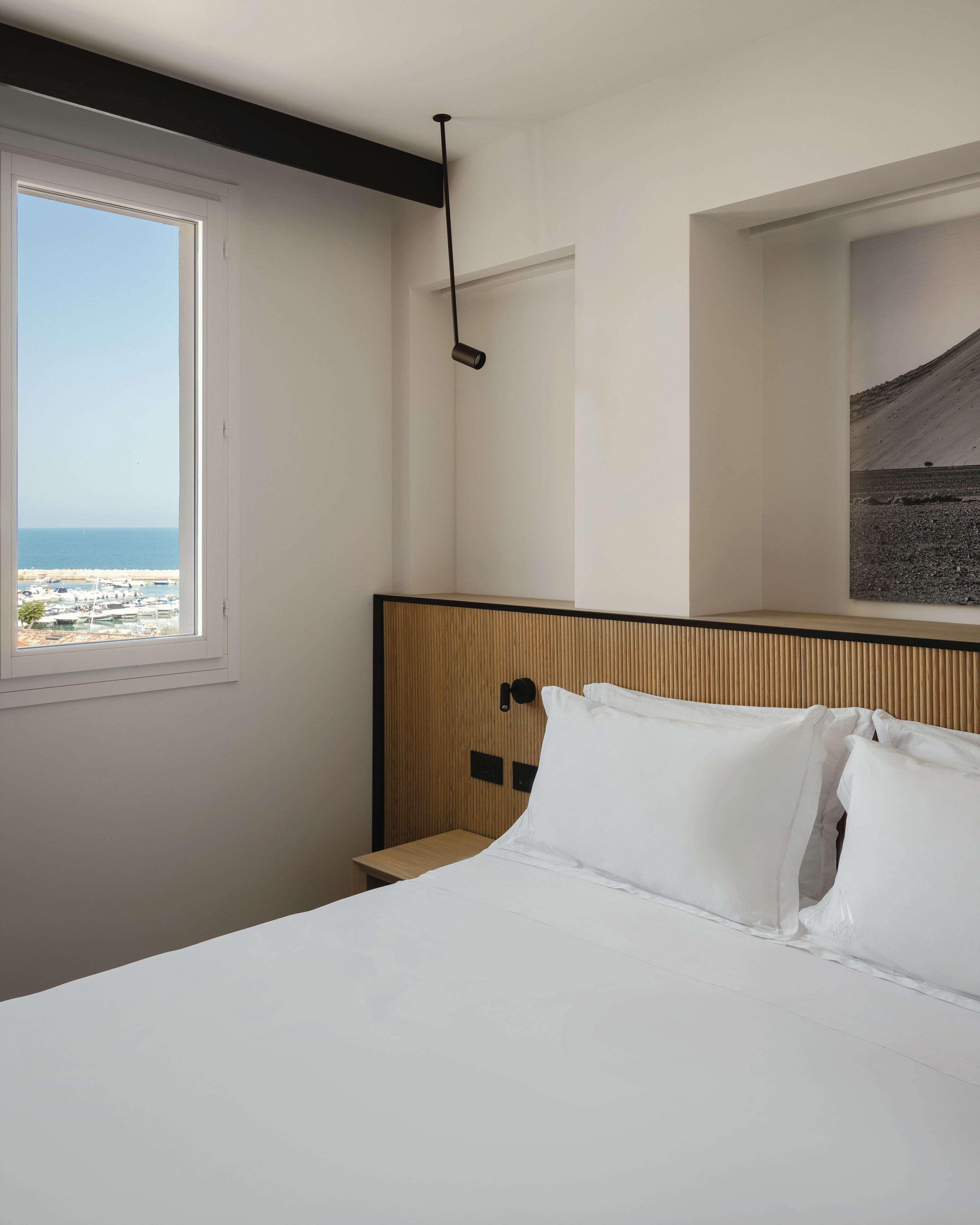 A hotel room with a bed, a nightstand, a wall-mounted lamp, a window with a view of the beach, and a framed picture on the wall.