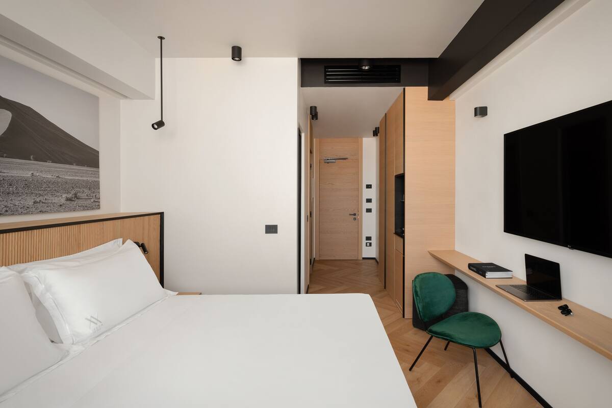 A modern hotel room with a large bed, a desk with a laptop, a green chair, and a TV mounted on the wall.