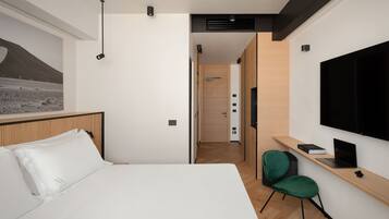 A modern hotel room with a large bed, a desk with a laptop, a green chair, and a TV mounted on the wall.