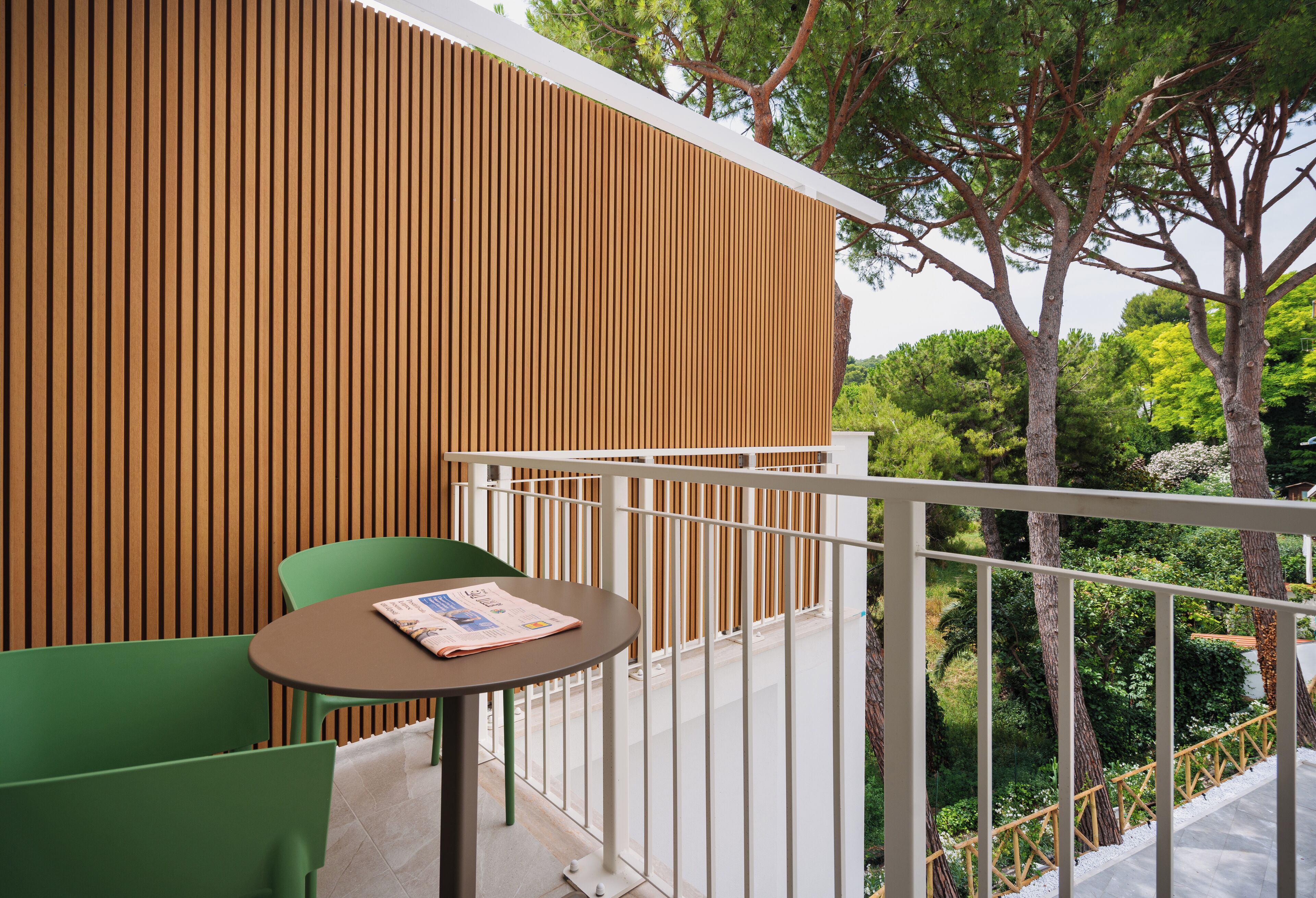 Un balcon doté d’un mur en lattes de bois, d’une chaise verte, d’une petite table ronde sur laquelle repose un journal, et d’une rambarde.