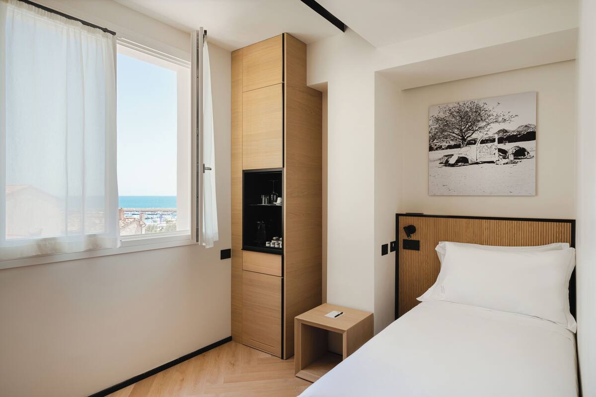 A hotel room with a bed, a small bedside table, a wardrobe, a window with curtains, and a wall-mounted artwork.