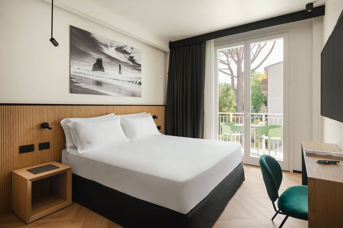 A modern hotel room with a large bed, a wooden headboard, a small bedside table, a desk with a chair, and a balcony with green chairs.