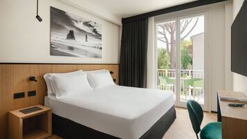 Una habitación de hotel moderna con una cama grande, un cabecero de madera, una mesita de noche pequeña, un escritorio con una silla y un balcón con sillas verdes.