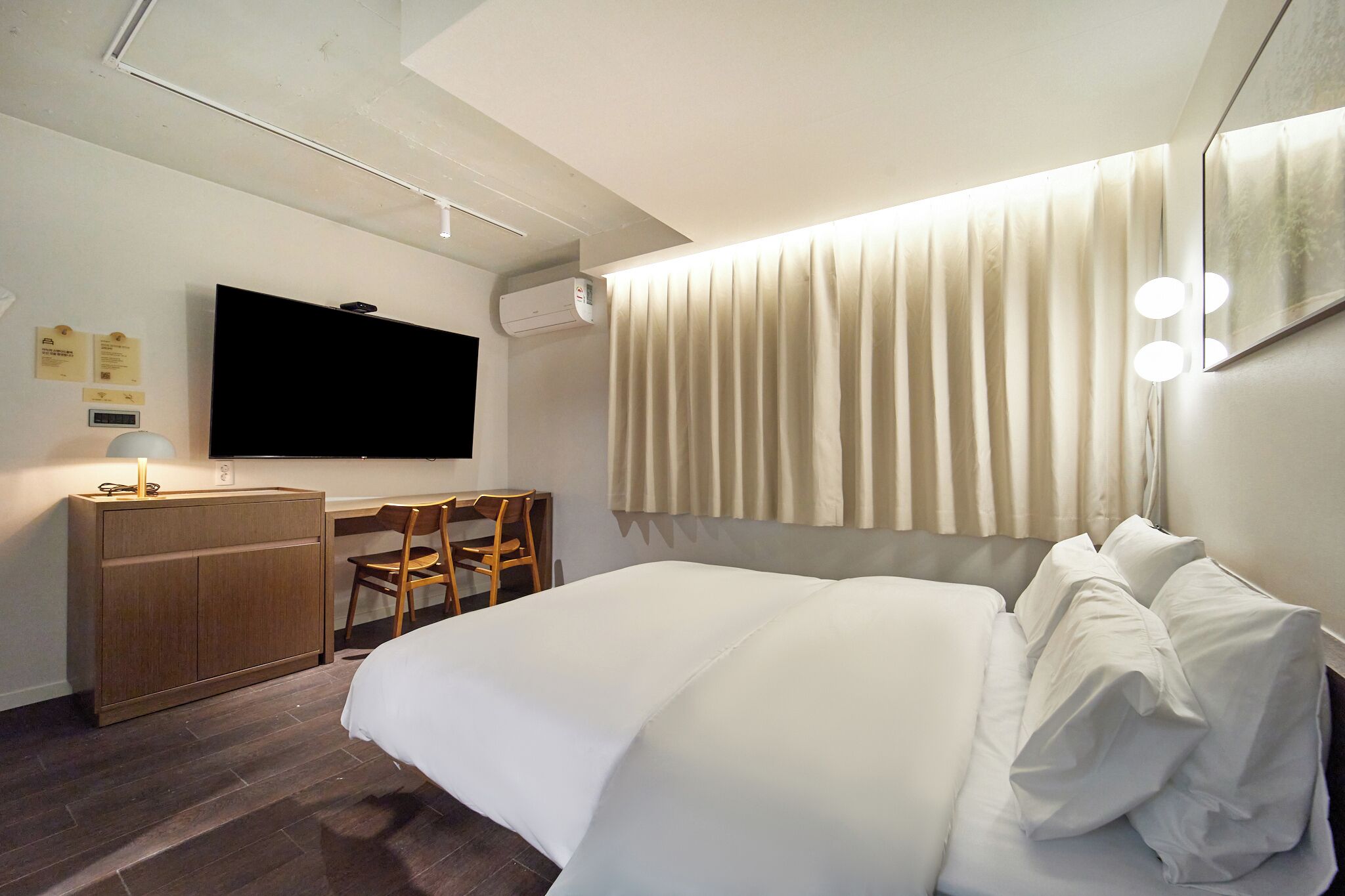 A hotel room with a bed, a desk with a chair, a flat-screen TV, and an air conditioning unit.