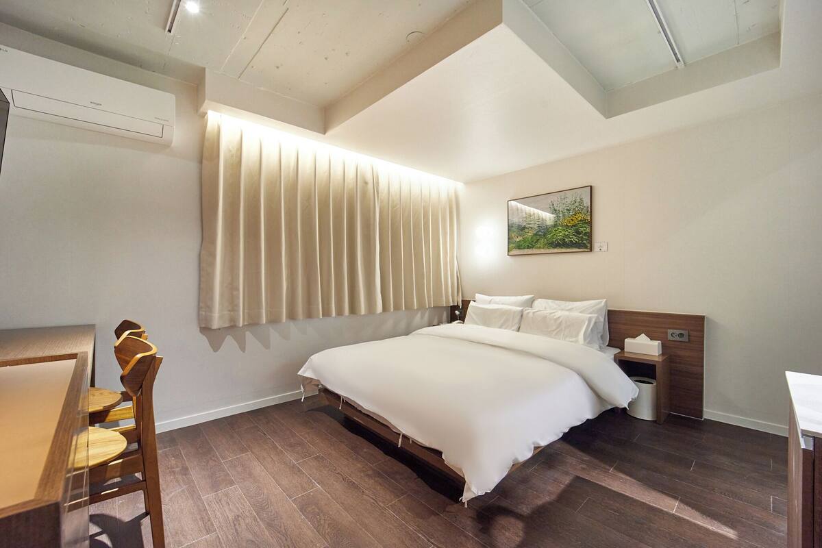 A bedroom with a bed, a bedside table, a nightstand, a window with curtains, a painting on the wall, and a wooden floor.