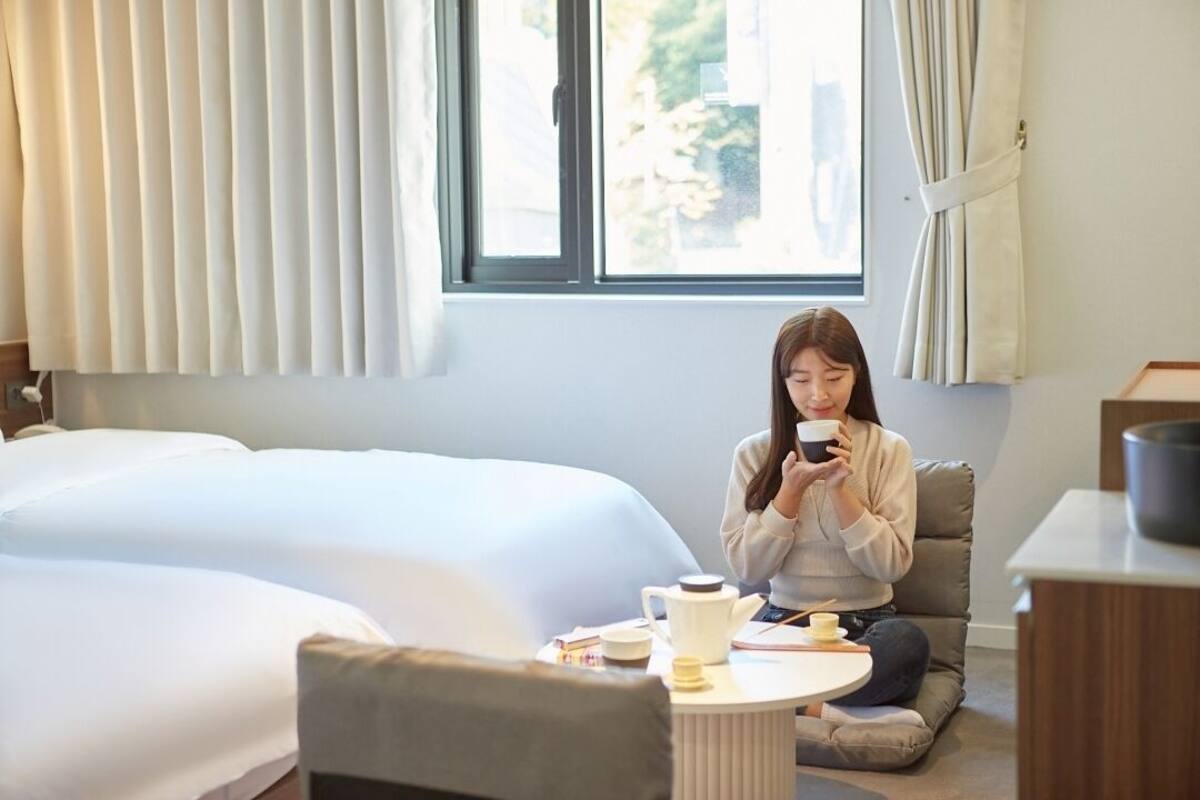 A hotel room with two beds, a small round table with a teapot and cups, and a person sitting on a couch using a smartphone.