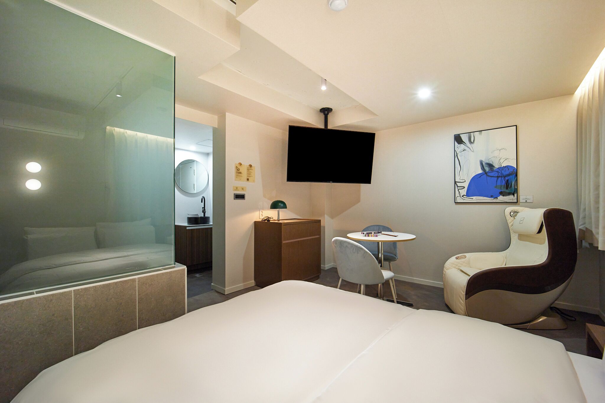 A modern hotel room with a large bed, a desk, a chair, a television, and a glass partition.