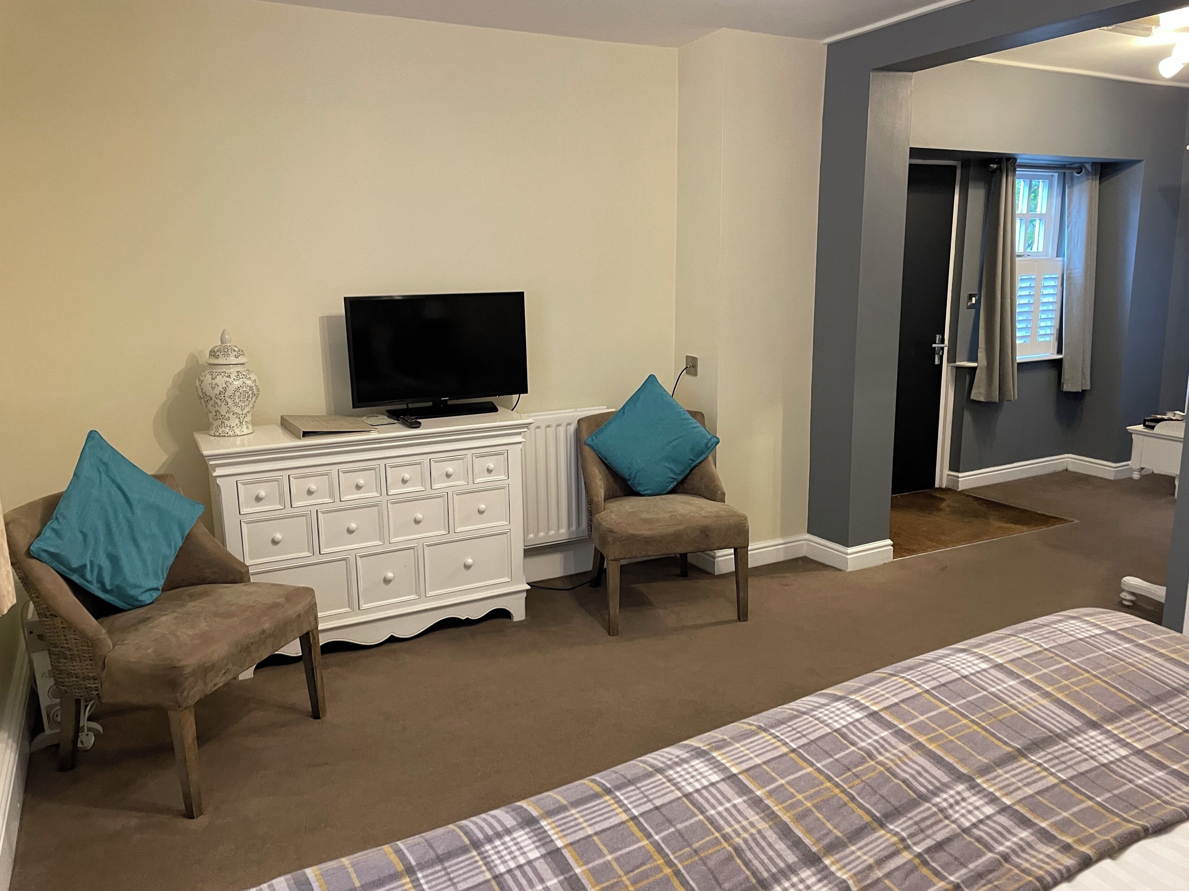 A bedroom with a bed, two chairs, a dresser, a television, and a door leading to a bathroom.