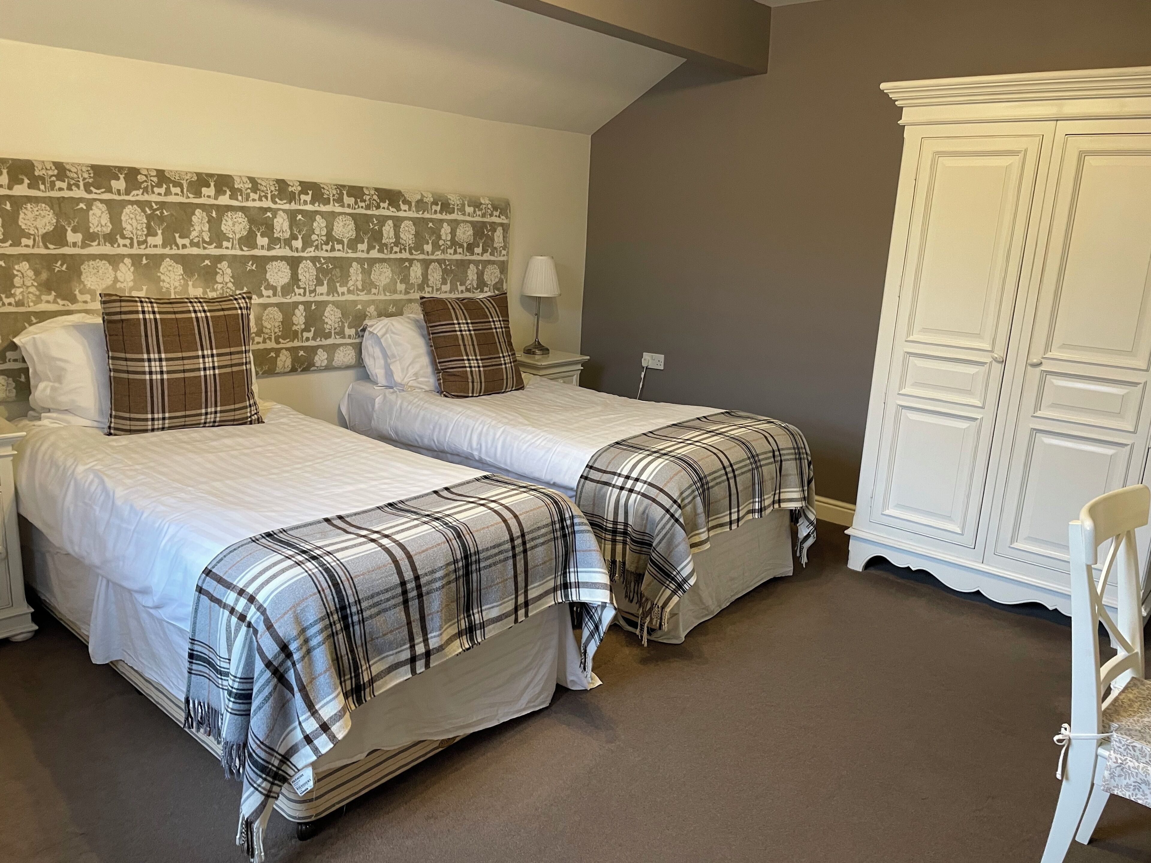 Ein Hotelzimmer mit zwei Betten, jedes mit einer Karo-Decke, einem Kopfteil mit Naturtapete, einem weißen Kleiderschrank und einem Stuhl.