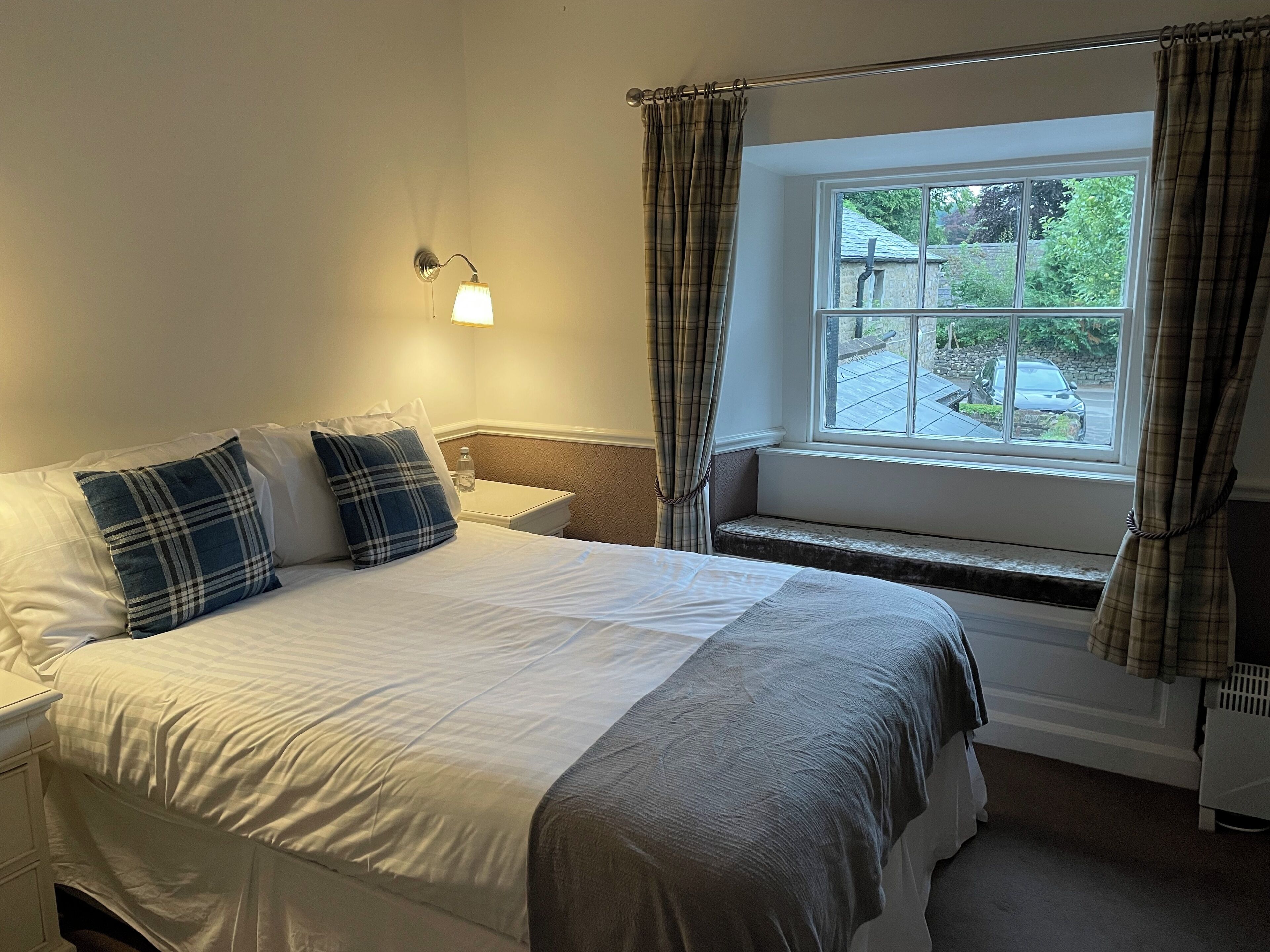 Een netjes opgemaakt bed met witte lakens en geruite kussens, een nachtkastje met een lamp en een raam met gordijnen.