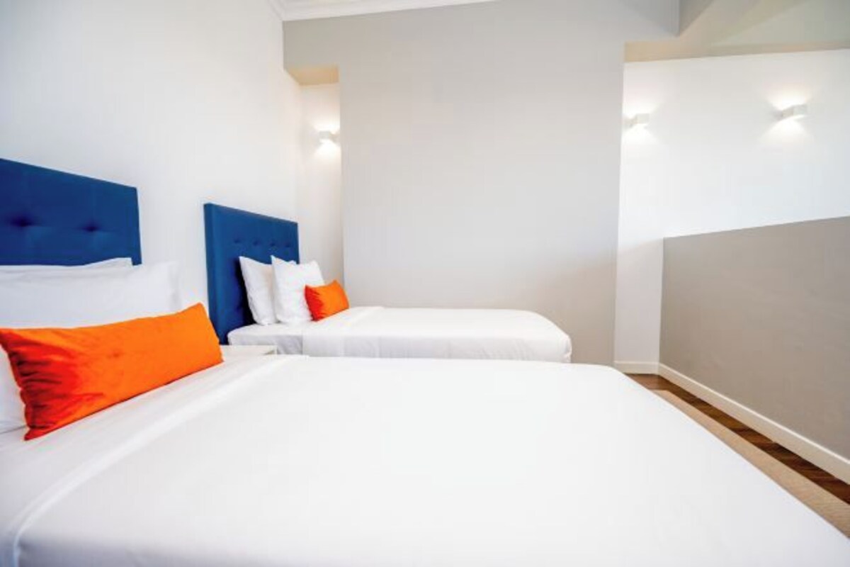 Habitación de hotel con dos camas, una con cabecera azul y la otra con cabecera blanca, ambas con ropa de cama blanca y almohadas anaranjadas.
