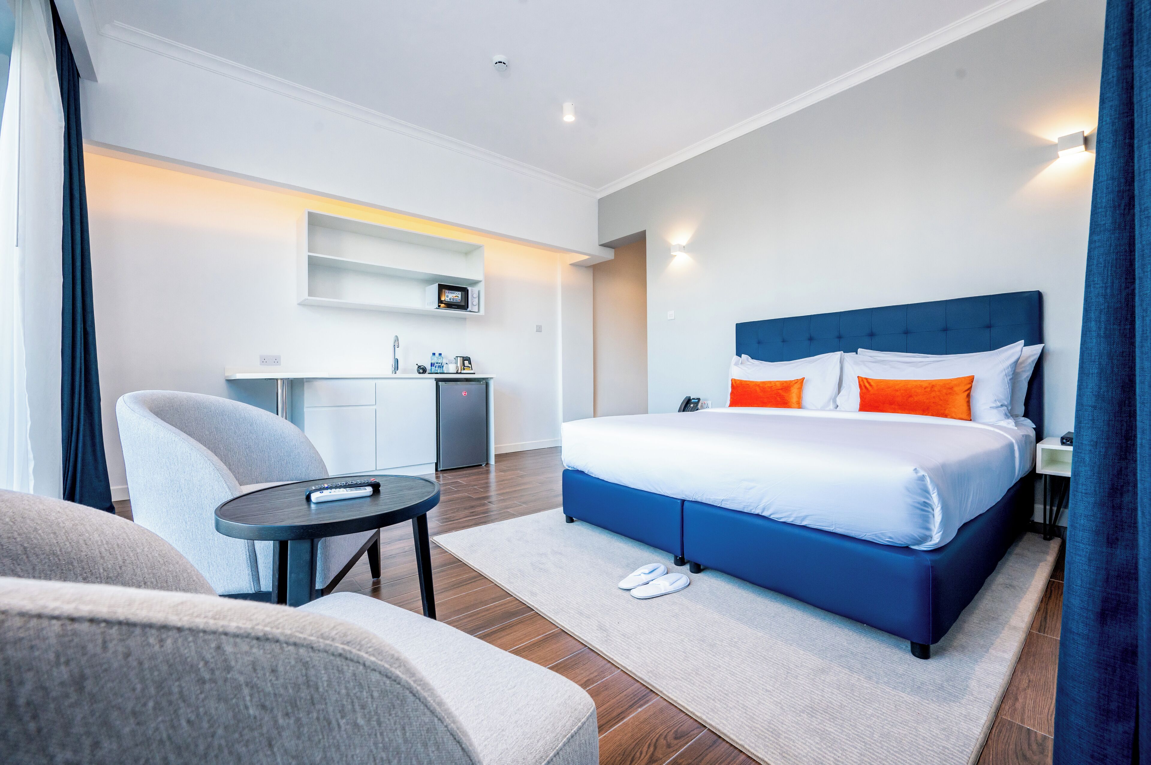A modern hotel room with a large bed, a small desk, a chair, and a kitchenette.