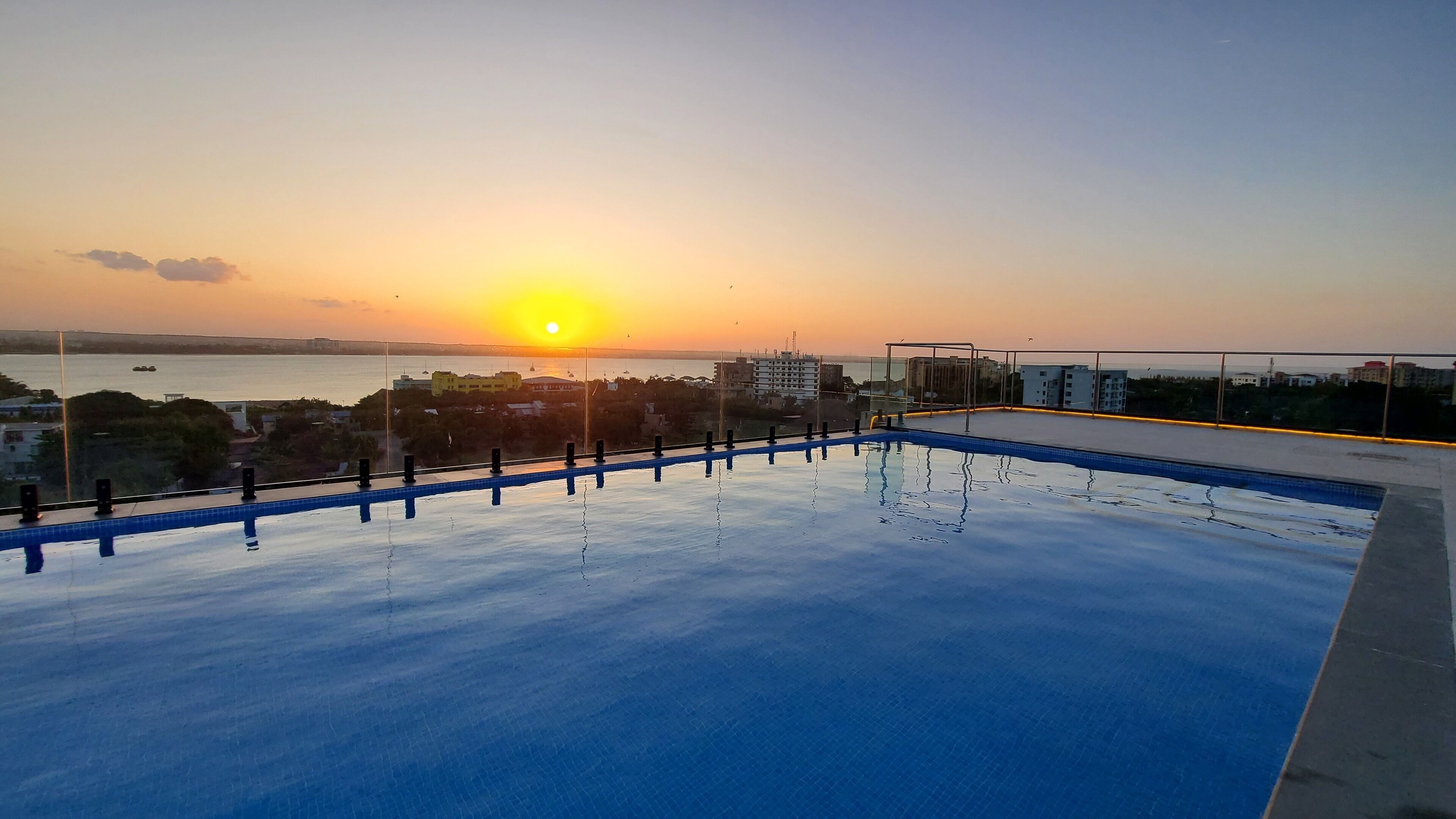 A rooftop pool with a view of the sunset over a body of water, surrounded by buildings.