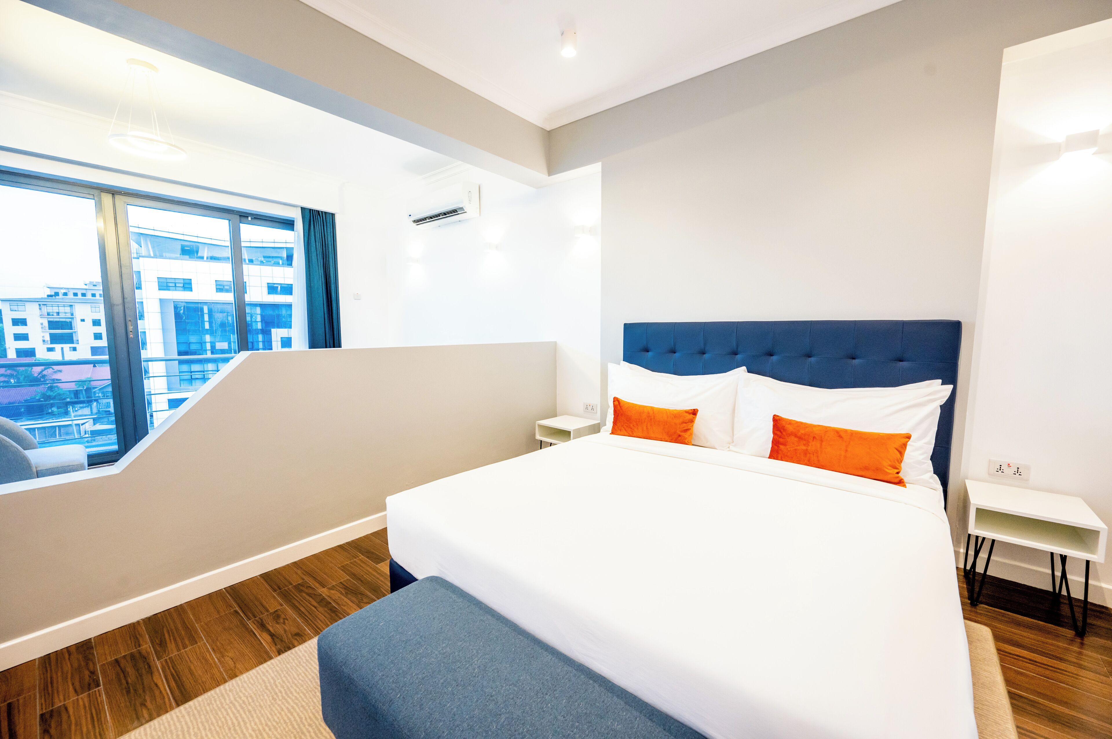 A modern hotel room with a large bed, a view of the city through floor-to-ceiling windows, a small bedside table, and a built-in bench at the foot of the bed.