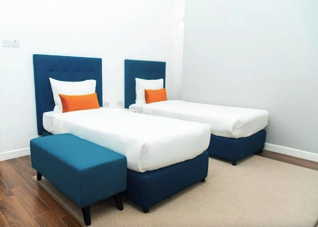 Une chambre avec deux lits simples, chacun doté d’une tête de lit bleue et de draps blancs, ainsi que de deux petits tabourets assortis de couleur bleue.
