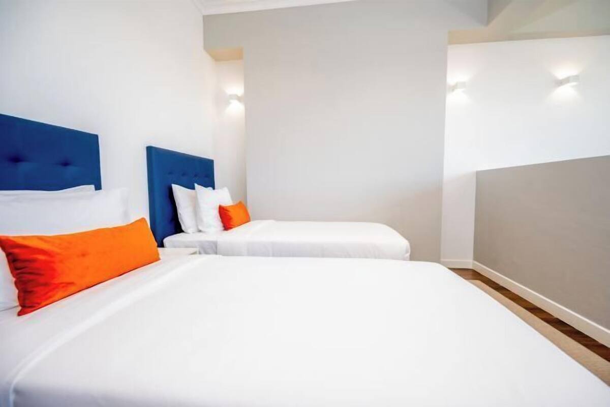 Ett hotellrum med två sängar, den ena med en blå sänggavel och den andra med en vit sänggavel. Båda sängarna har vita sängkläder och orange kuddar.
