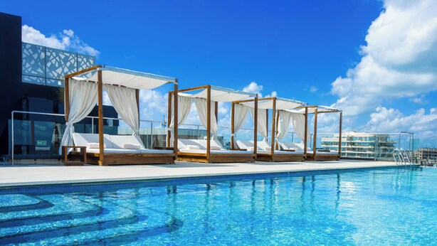 A poolside area with four wooden beds with white canopies, a clear blue pool, and a modern building in the background. - Urban by CityBlue Hotel, Dar es Salaam (Dar es Salaam)