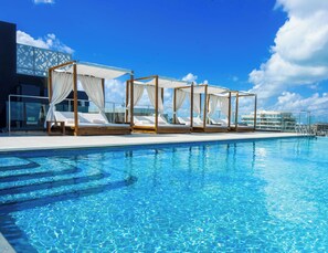 A poolside area with four wooden beds with white canopies, a clear blue pool, and a modern building in the background. - Urban by CityBlue Hotel, Dar es Salaam (Dar es Salaam)