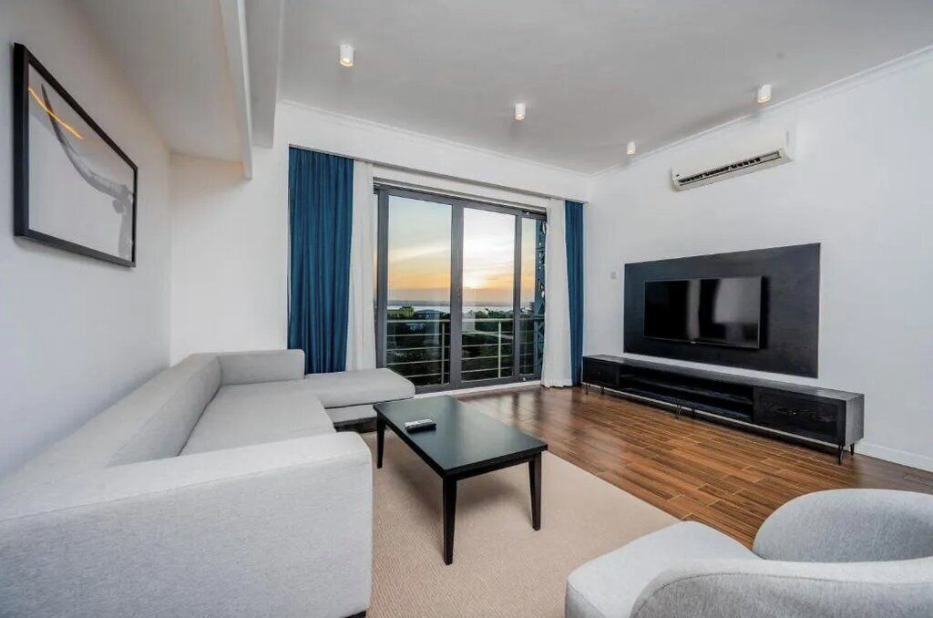 A modern living room with a large flat-screen TV, a sofa, a coffee table, and a view of the city through large windows.